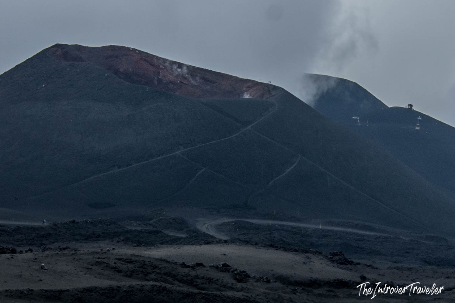 Mount Etna summit craters with volcanic steam, red lava rock, and hiking trails on black ash slopes