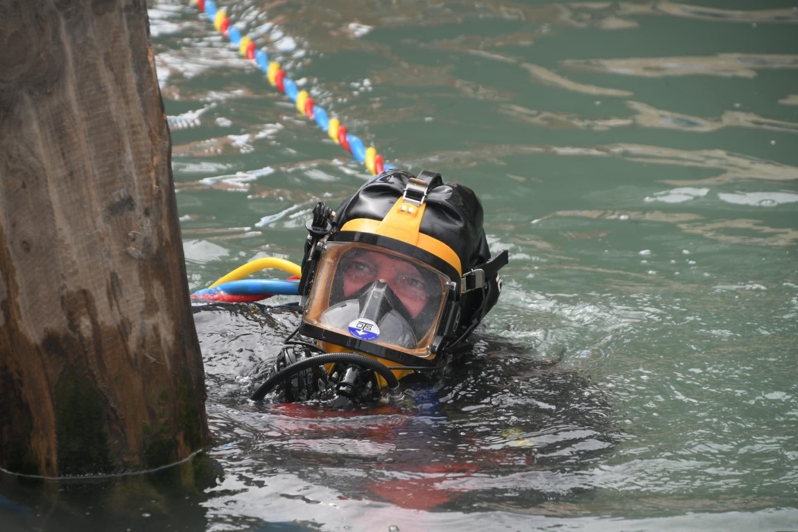 Professional venice canal diver surfacing near a wooden piling wearing an OTS full-face diving helmet