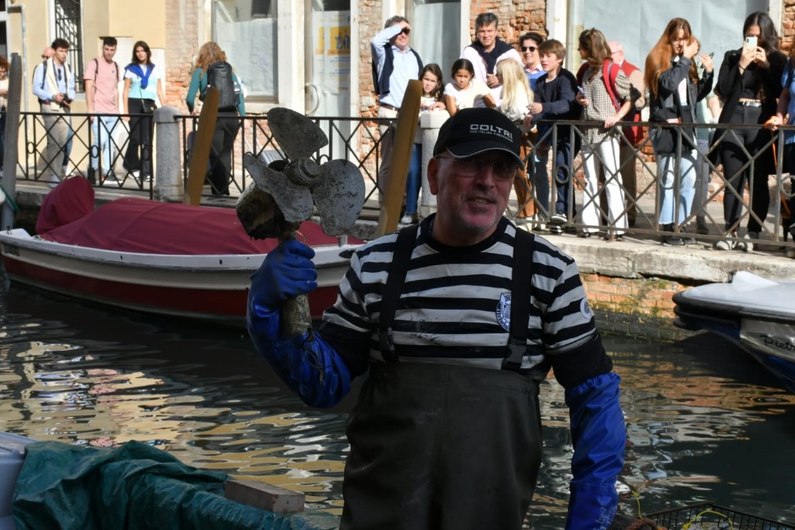 Venetian canal diver holding a retrieved boat propeller beside a Venice waterway, watched by onlookers