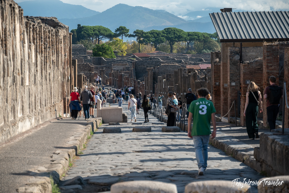 Tourists walking along an ancient cobblestone street in Pompeii, with ruined brick walls and mountains beyond