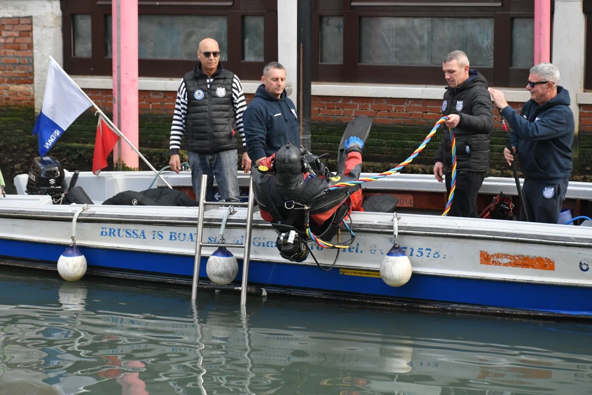 Scuba diver entering a Venice canal from a Brussa Is Boat vessel, assisted by crew members on deck