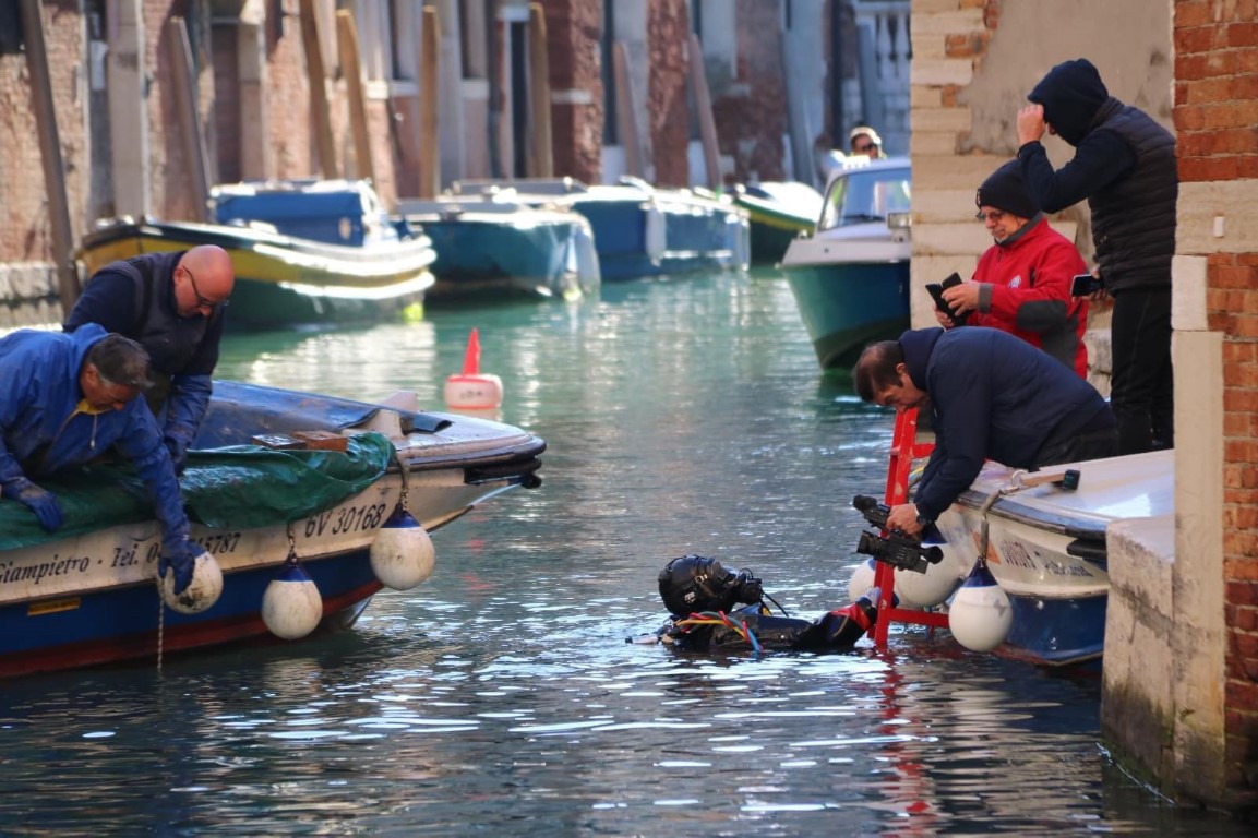 Scuba diver with camera equipment submerged in a Venice canal, assisted by crew on moored boats