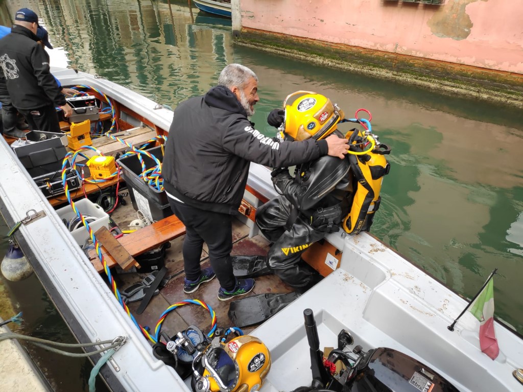 Technician helping a professional diver in Viking dry suit prepare on a boat in a Venice canal