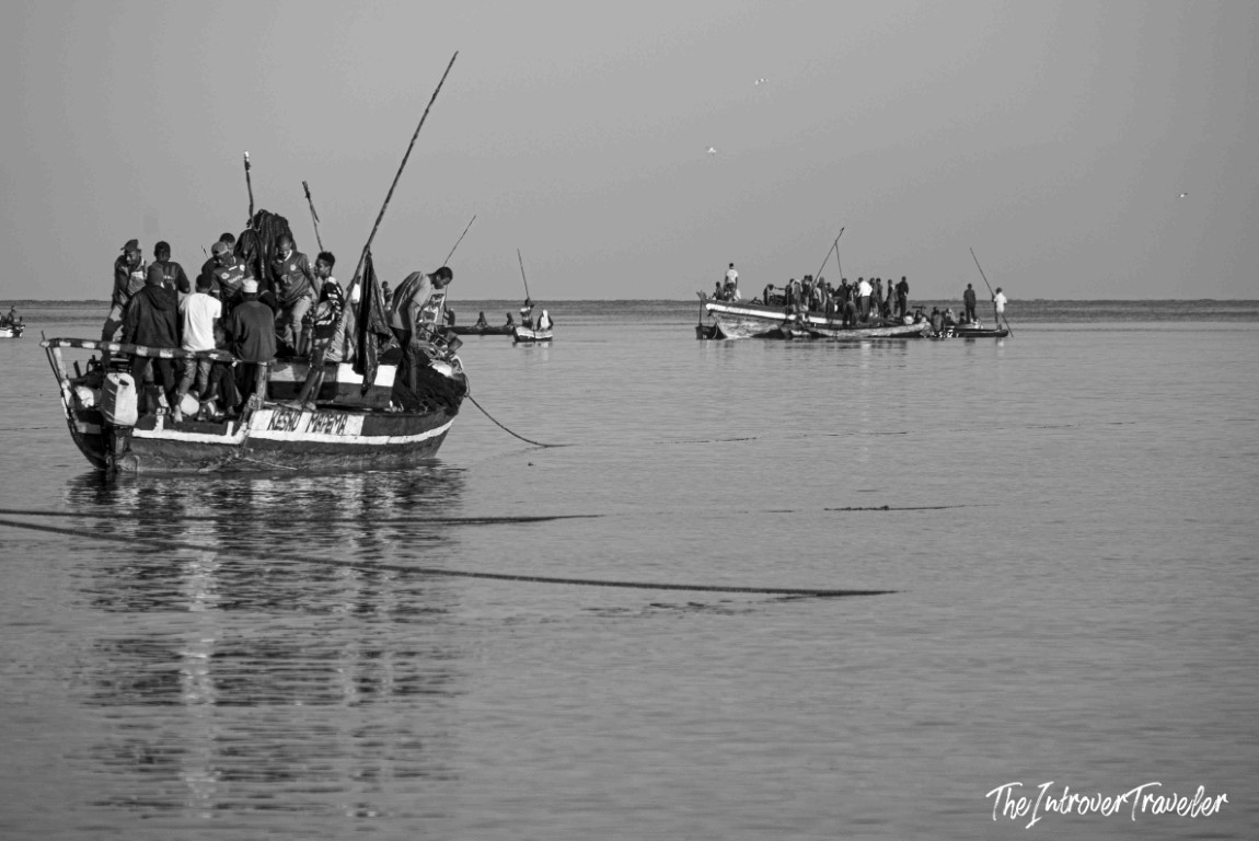 Black and white scene of crowded fishing boats at a coastal fish market, nets trailing in calm water