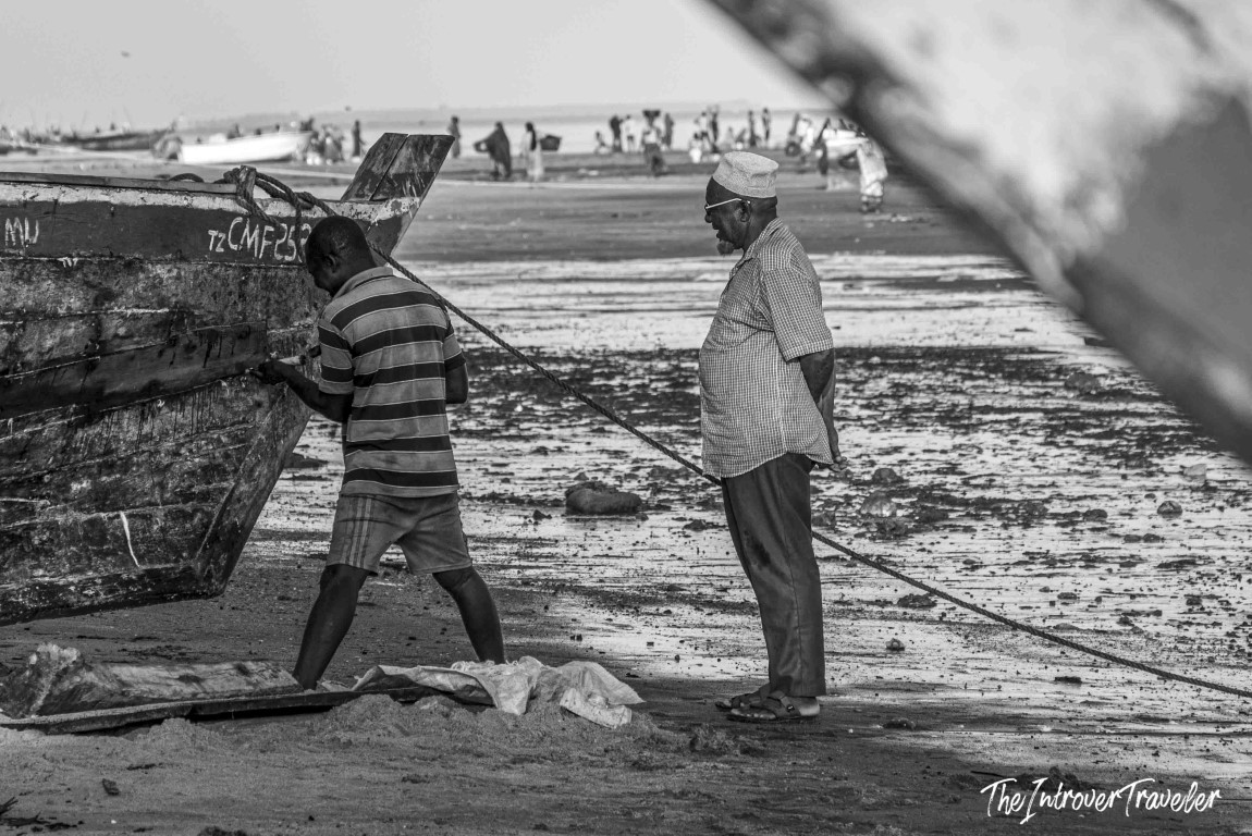Two Tanzanian fishermen hauling a wooden boat ashore at a busy coastal fish market beach