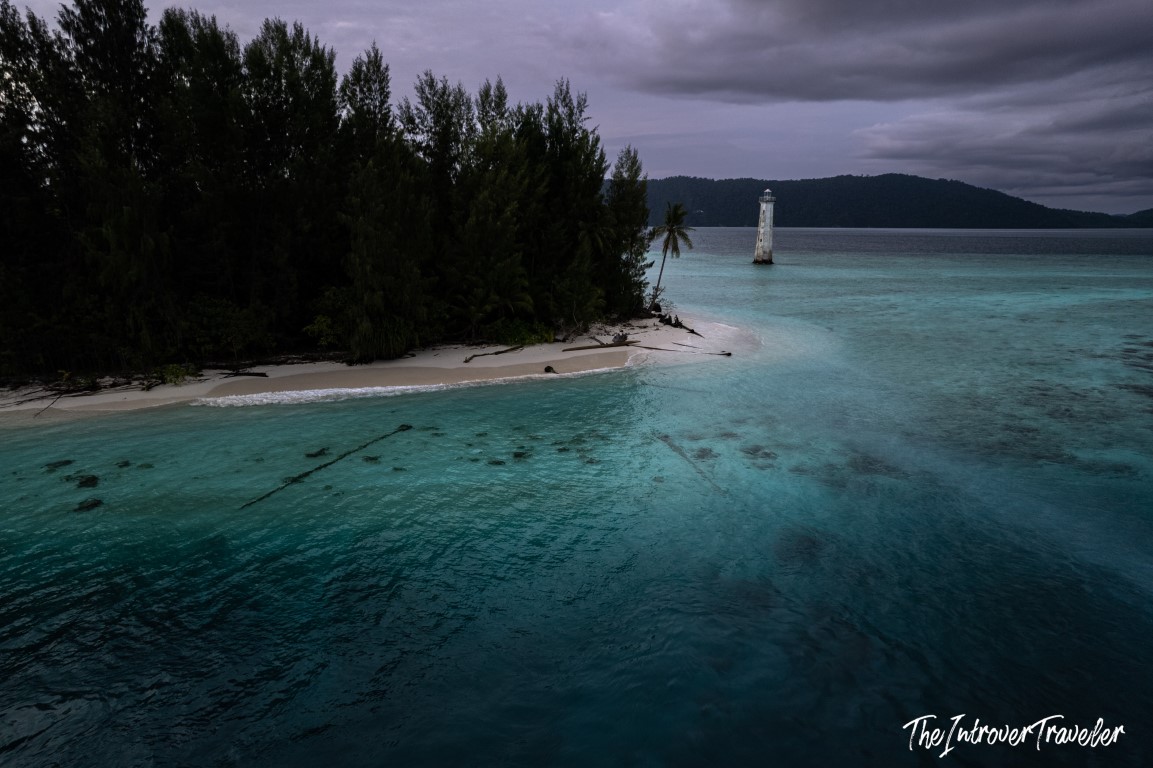 Aerial view of a remote Raja Ampat island tip, with turquoise reef waters and a lone lighthouse offshore