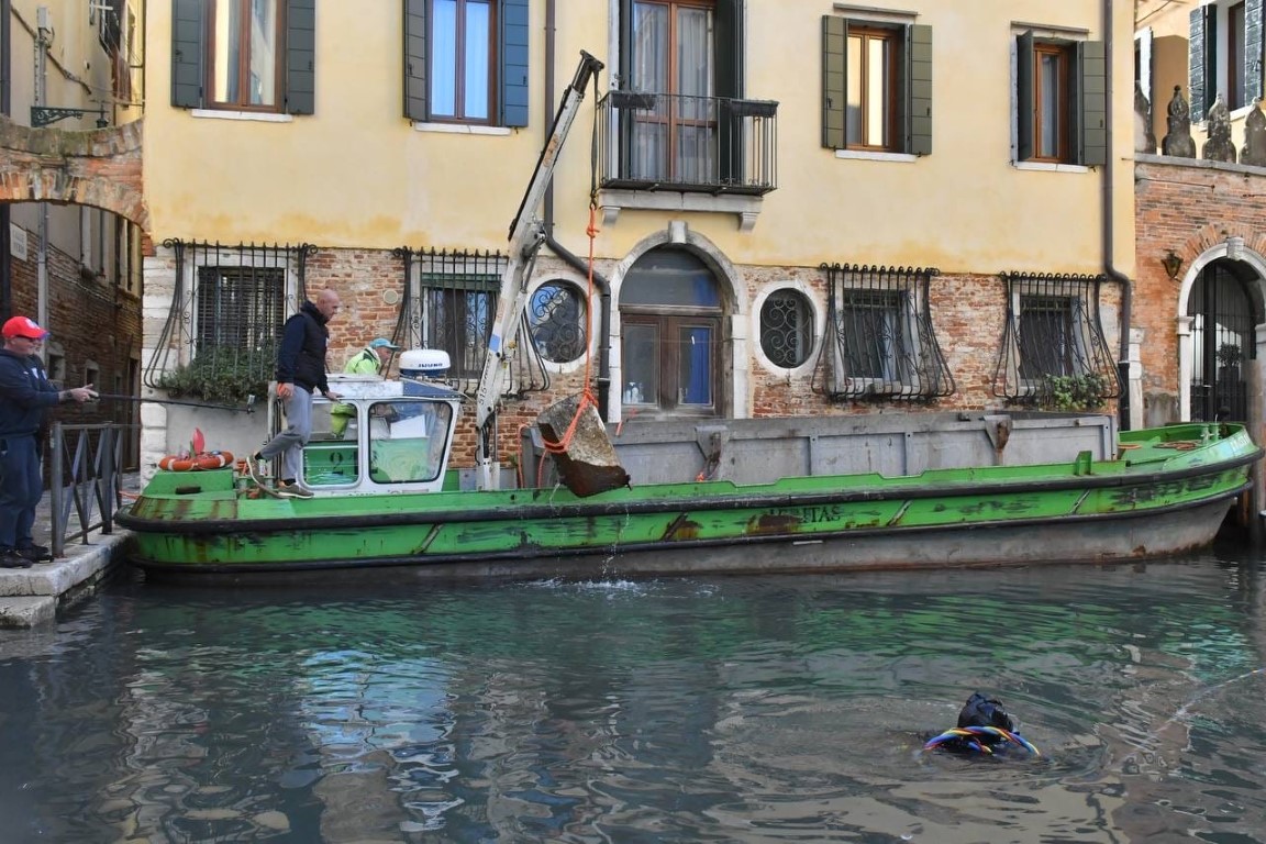 Venice canal maintenance crew lifting debris with crane barge as diver surfaces in narrow canal