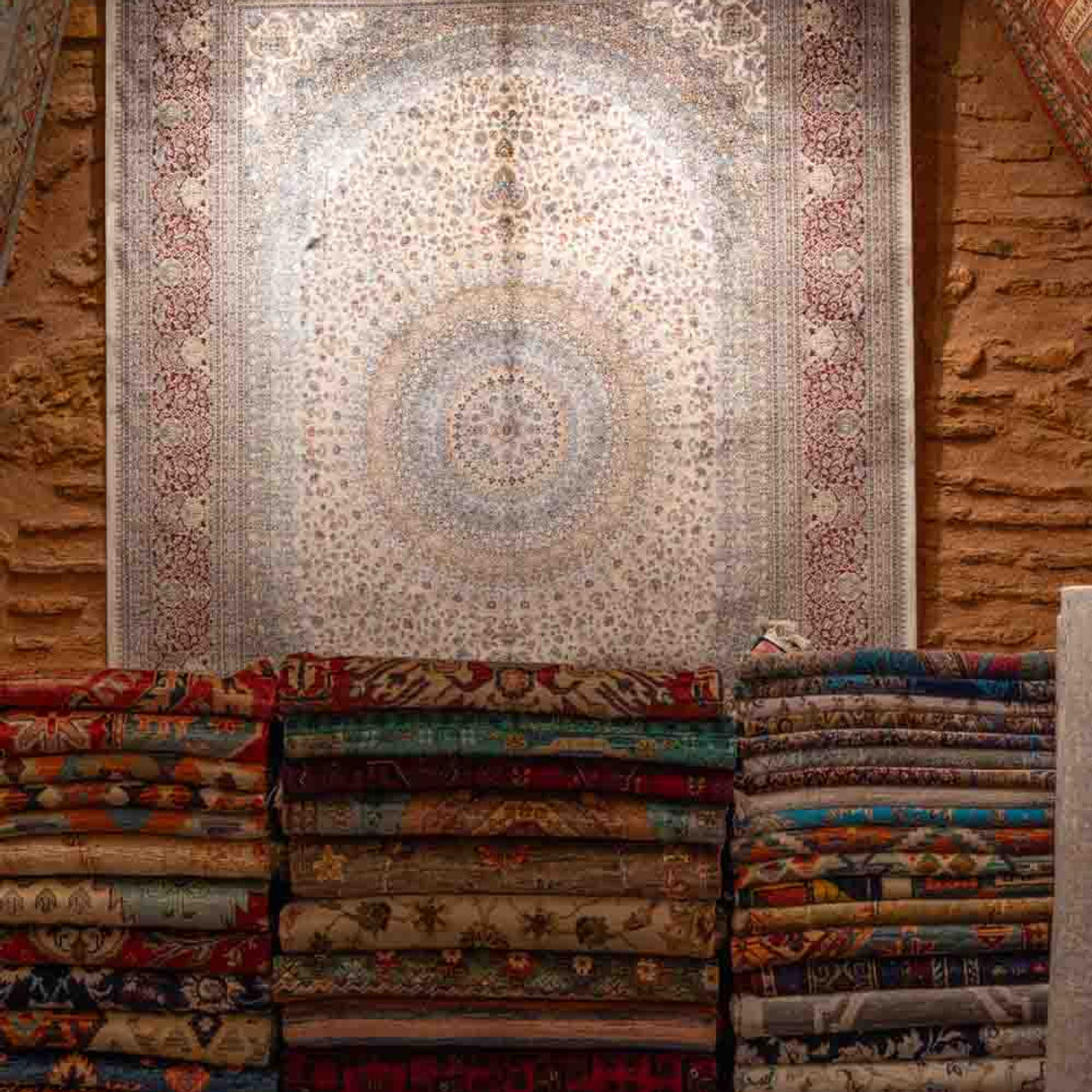 Stacked Persian-style rugs for sale in an Istanbul carpet shop, with ornate medallion rug displayed on stone wall
