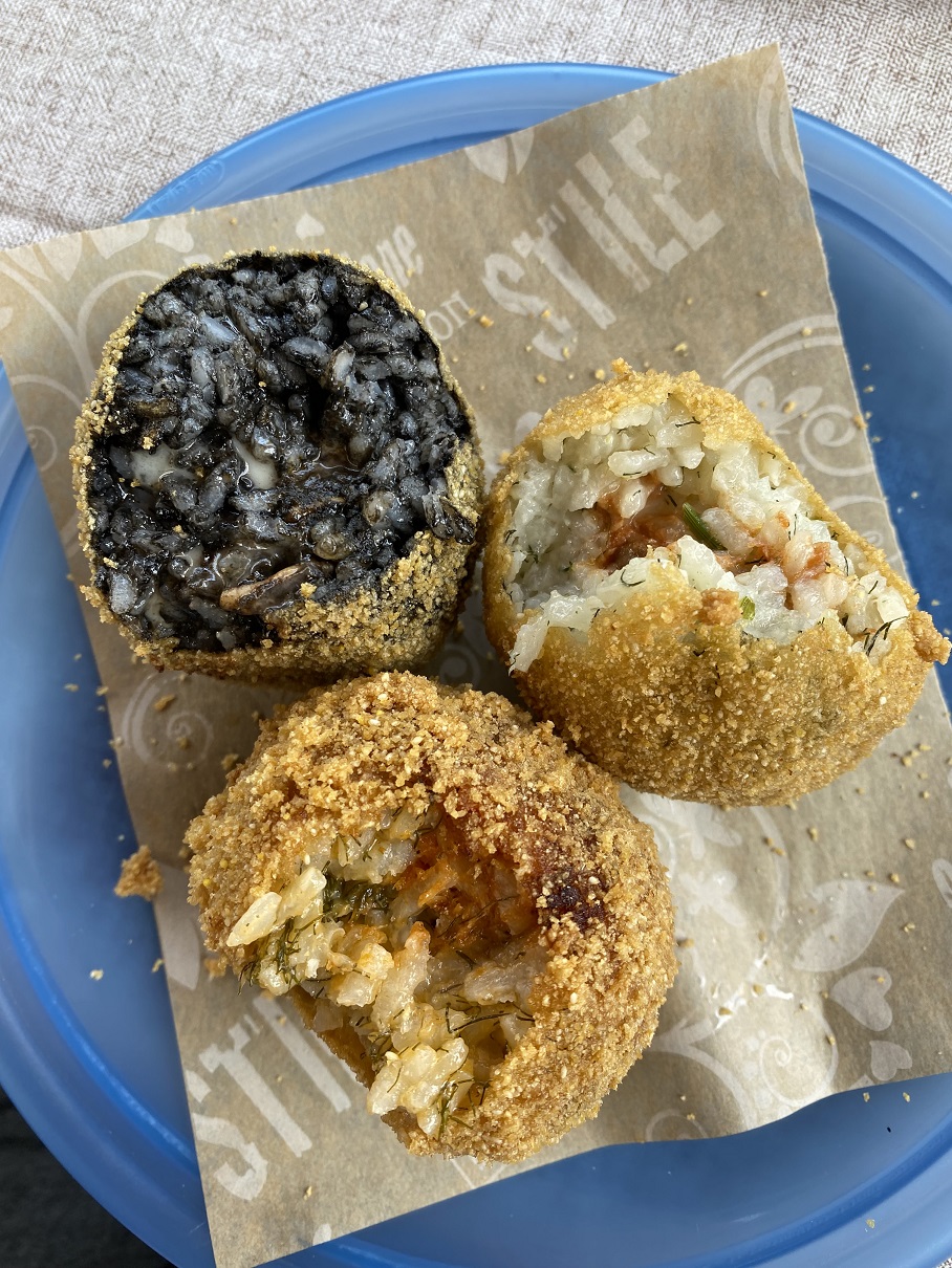 Three Sicilian arancini on a plate, one filled with squid ink rice, two others sliced open revealing savory fillings