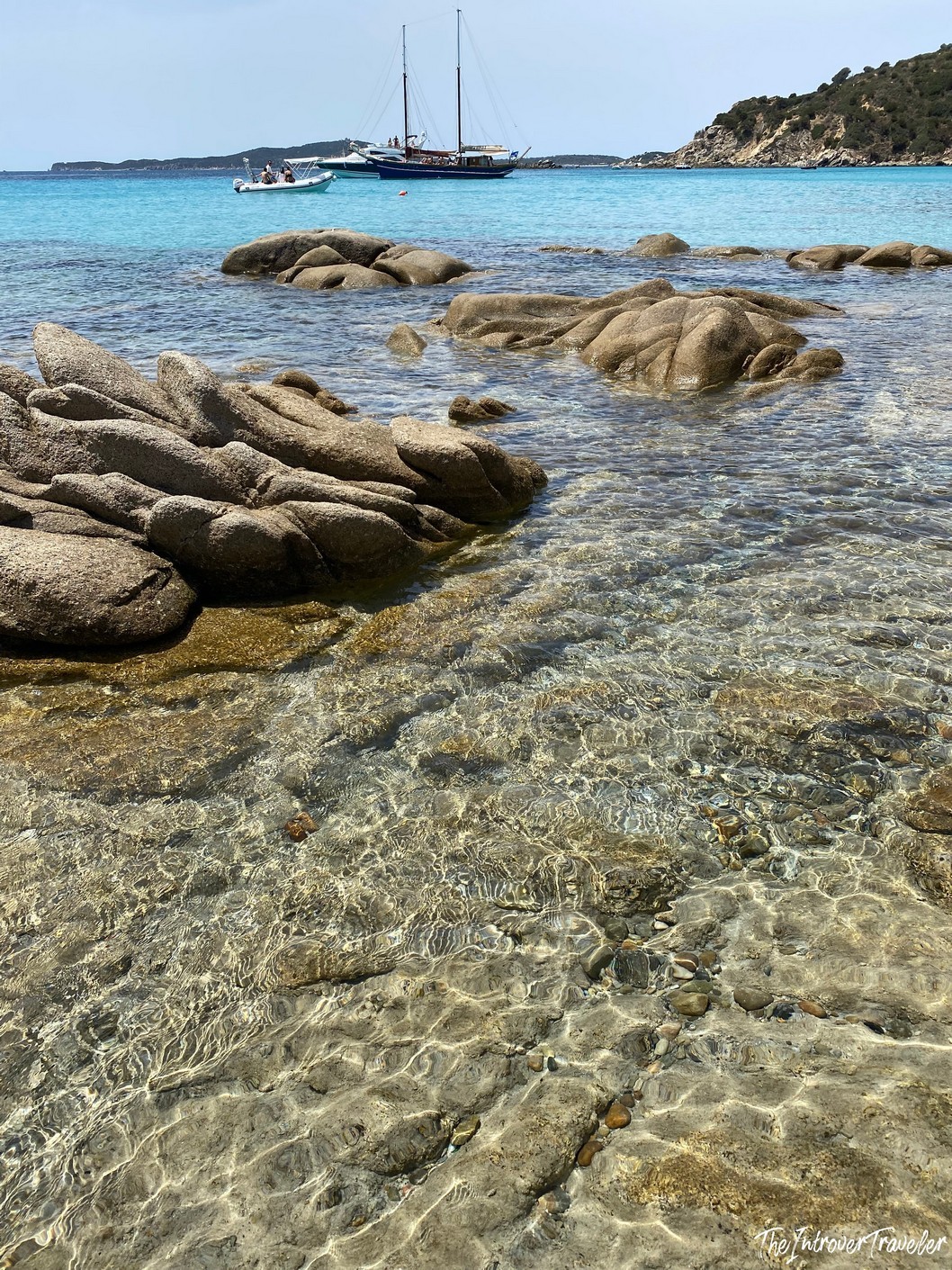 Crystal-clear shallow water over pebbles with granite boulders and a sailboat anchored in turquoise sea, south Sardinia