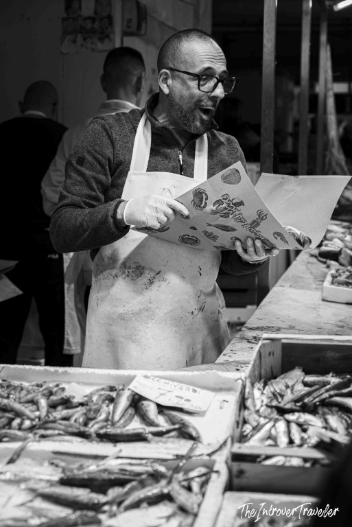 Animated fish vendor holding a seafood menu at a bustling coastal city fish market