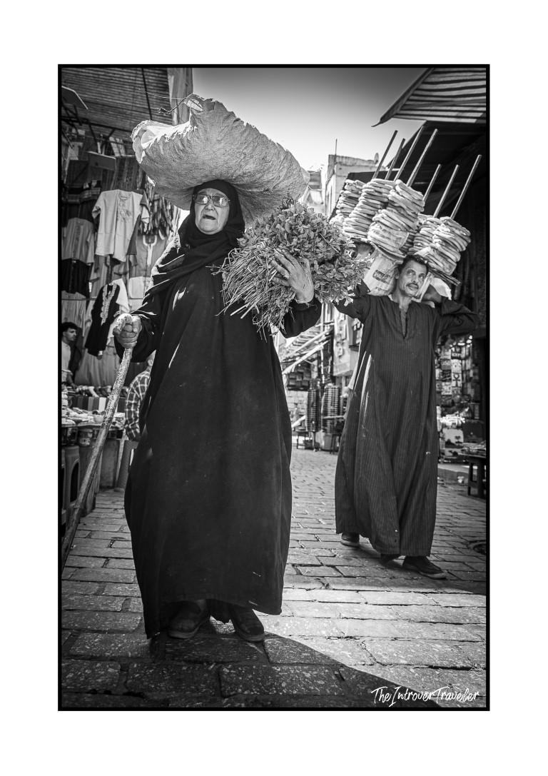 Black and white street scene in Egypt, woman carrying herbs and a man balancing stacked bread in a busy souk