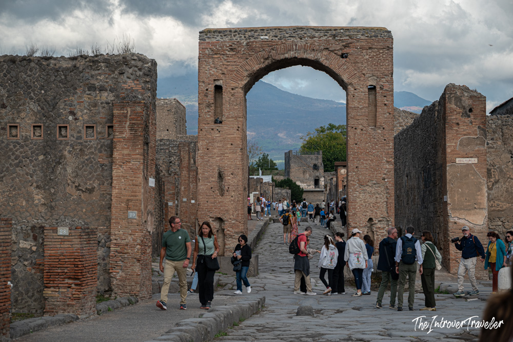 Tourists walking through the Arch of Caligula on a cobblestone street in Pompeii, with Mount Vesuvius behind