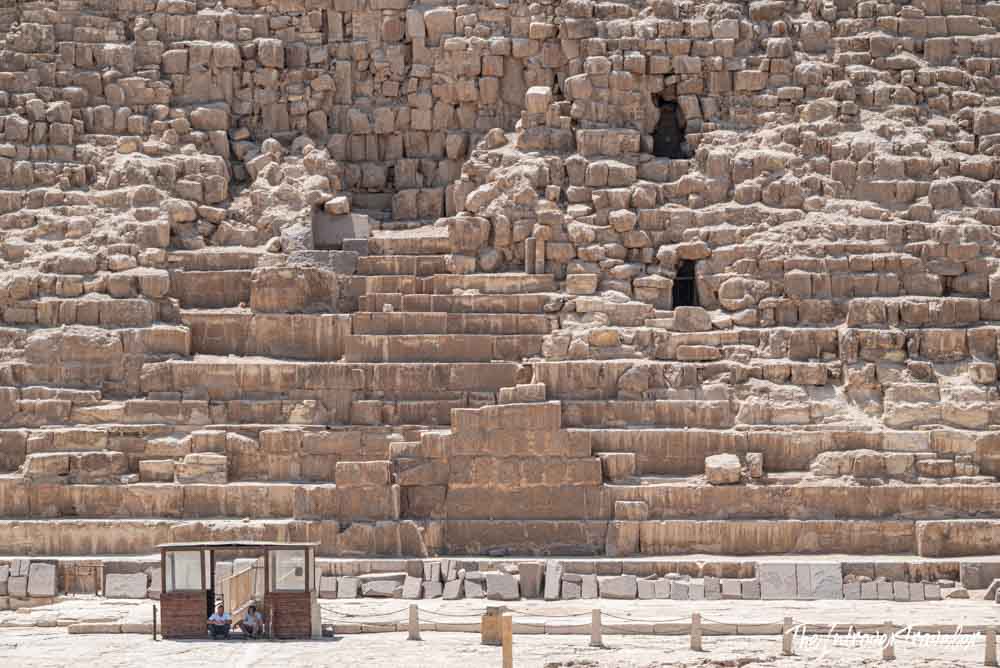 Close-up of massive limestone blocks forming the base of a Giza pyramid, with a guard post below showing the true scale.