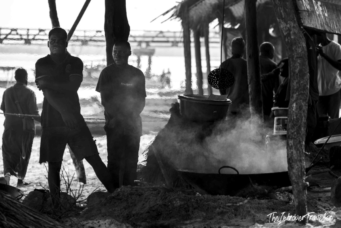 Men cooking over steaming pots at a coastal beach fish market, with ocean and pier in background