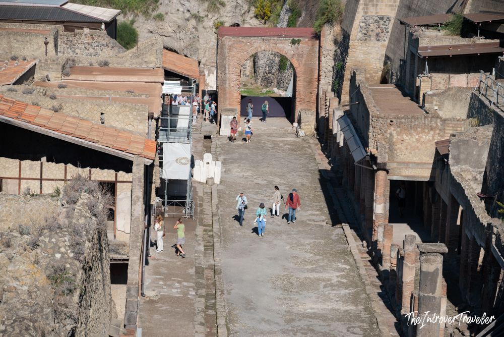 Aerial view of Herculaneum excavations, showing ancient Roman streets and a brick arch gate with visitors exploring below.