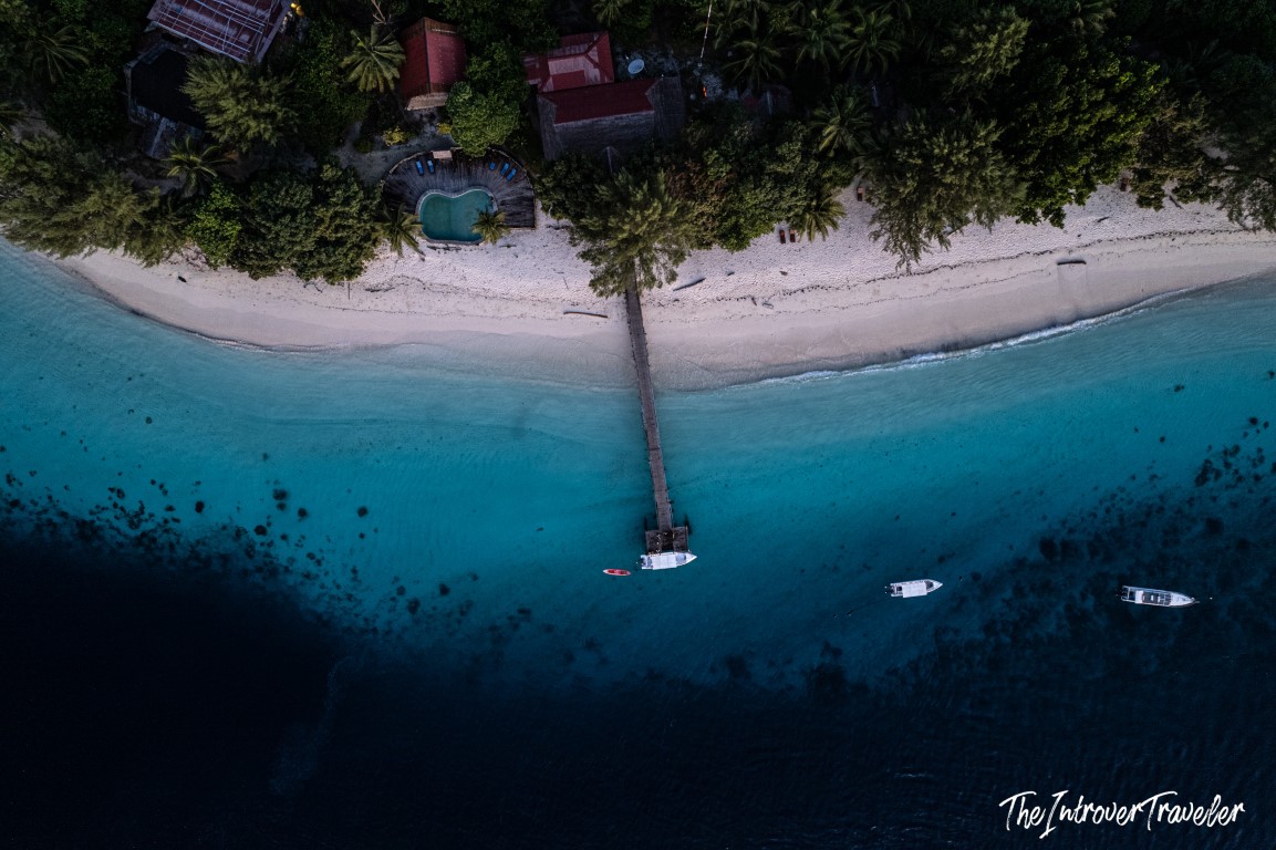 Aerial drone view of a Raja Ampat island resort, with a wooden jetty, white sand beach, and turquoise coral-rich waters.