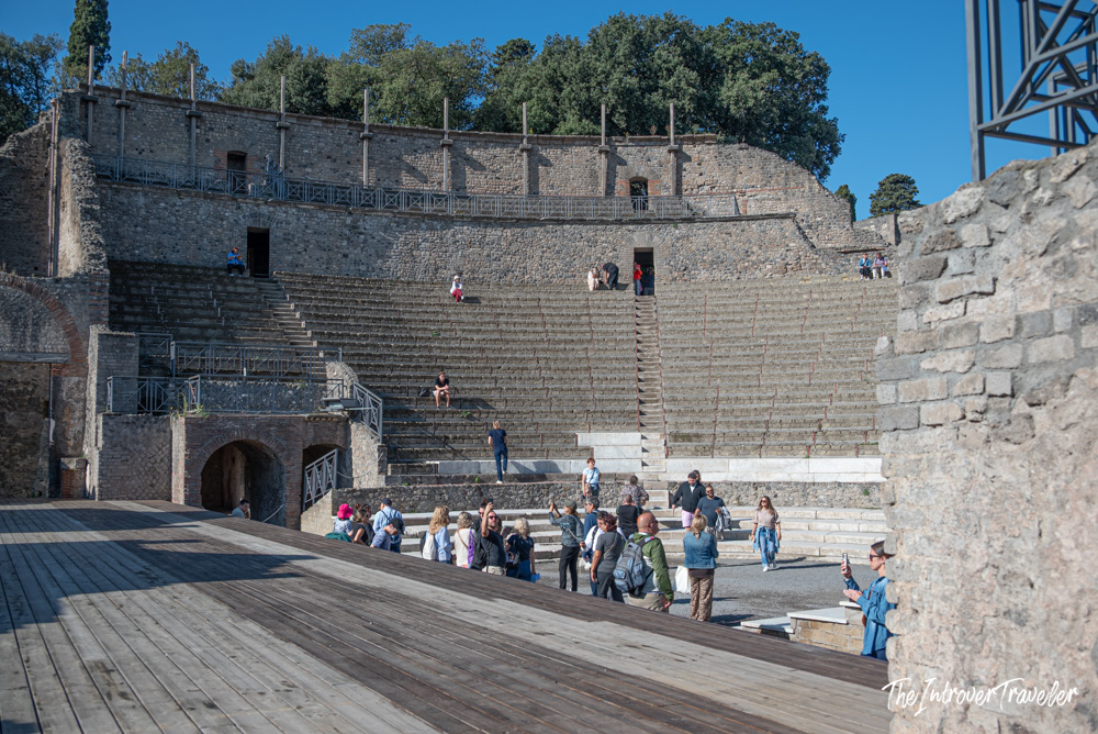 Tourists exploring the ancient Roman theatre at Pompeii, with tiered stone seating and wooden stage floor