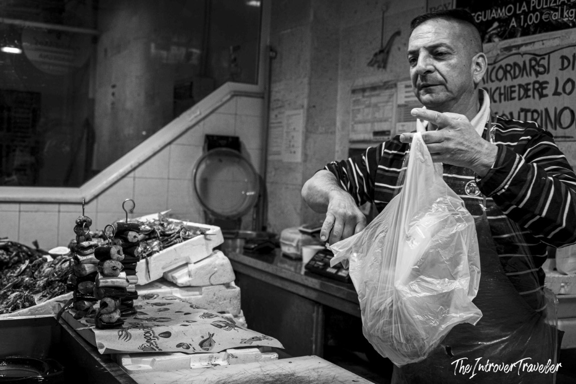 Italian fish market vendor packing a seafood purchase, with skewered shellfish and a market scale behind him