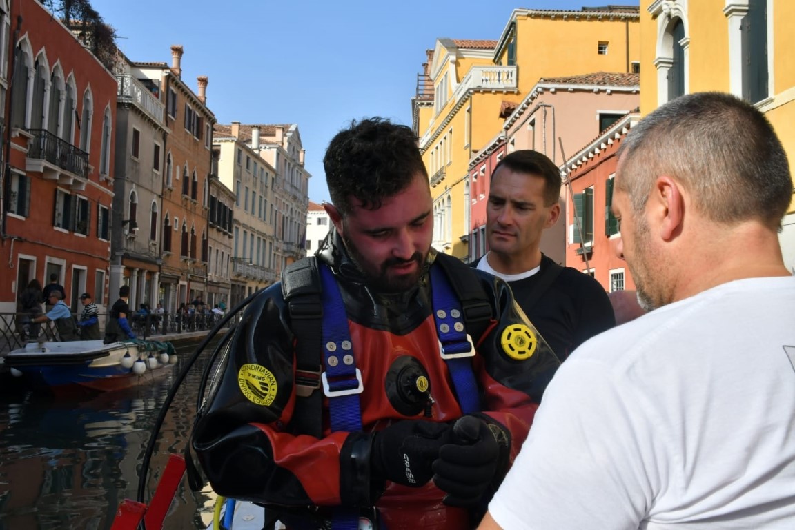 Diver in dry suit preparing to enter a Venice canal, flanked by two crew members on the canalside