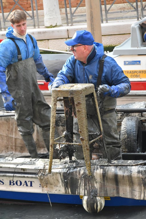 Workers in waders retrieving a mud-encrusted stool from a Venice canal cleanup boat