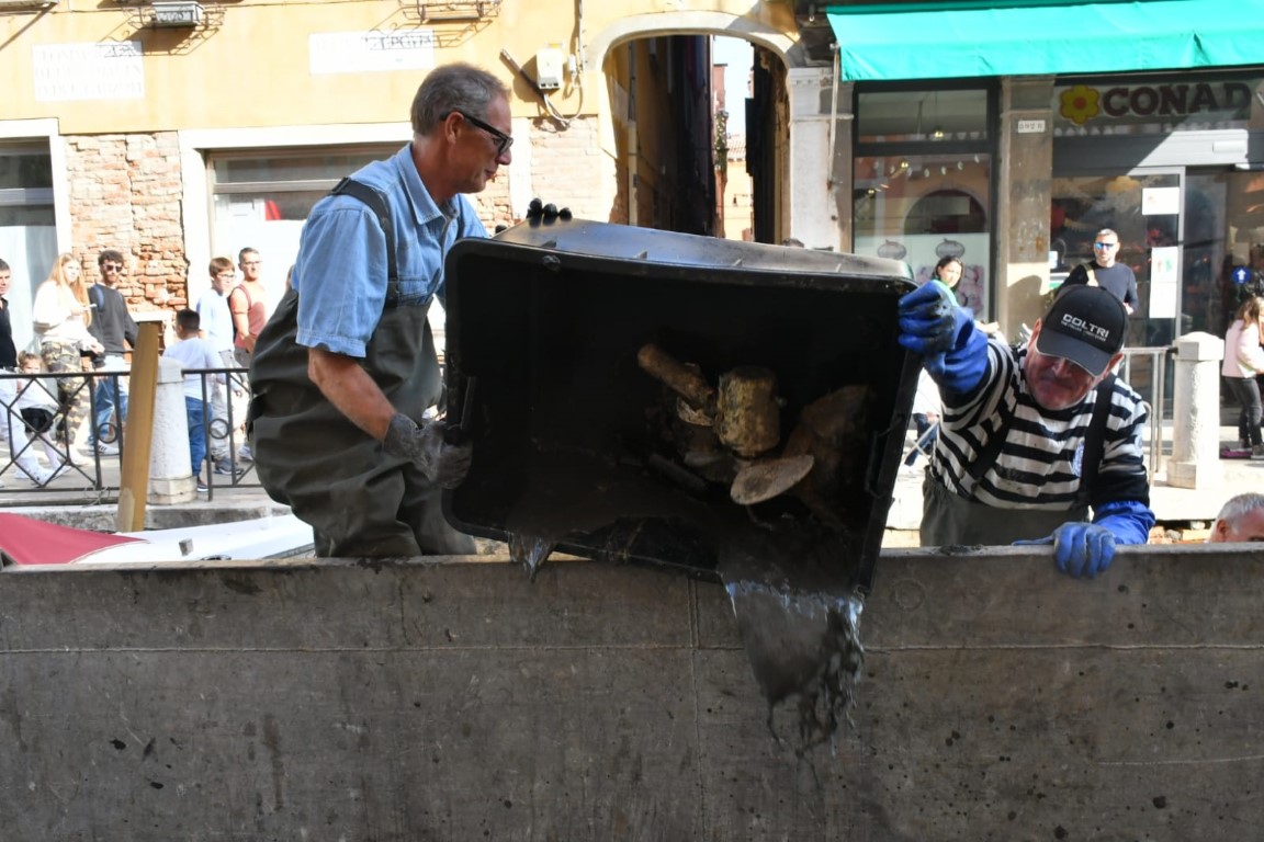 Two workers in waders removing debris from a Venice canal during a cleanup operation
