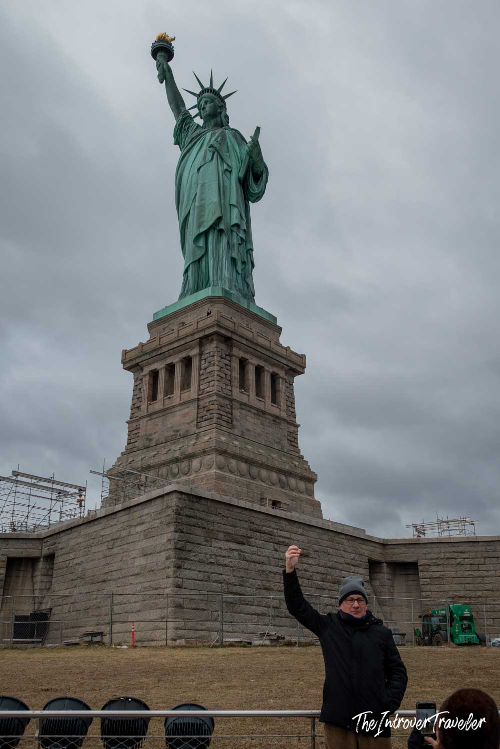 Visitor posing with raised arm in front of the Statue of Liberty on Liberty Island, overcast sky