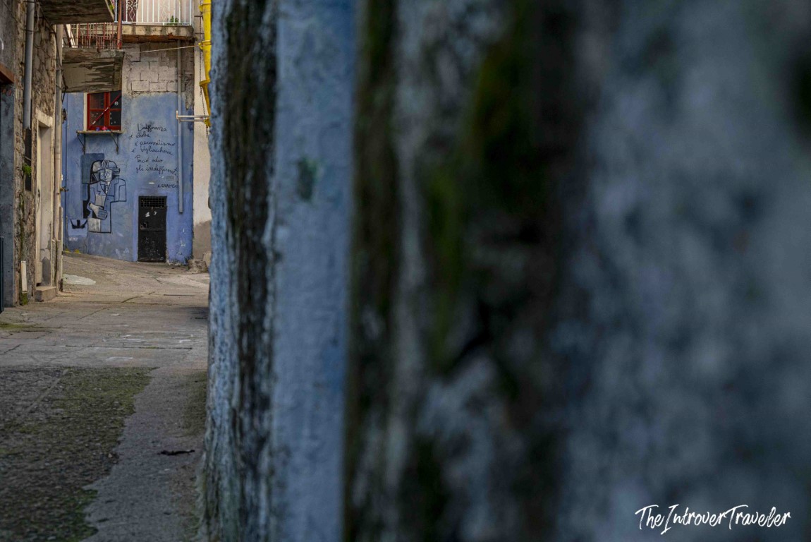 Narrow alley in Orgosolo framing a blue wall mural with Italian political text and a painted figure