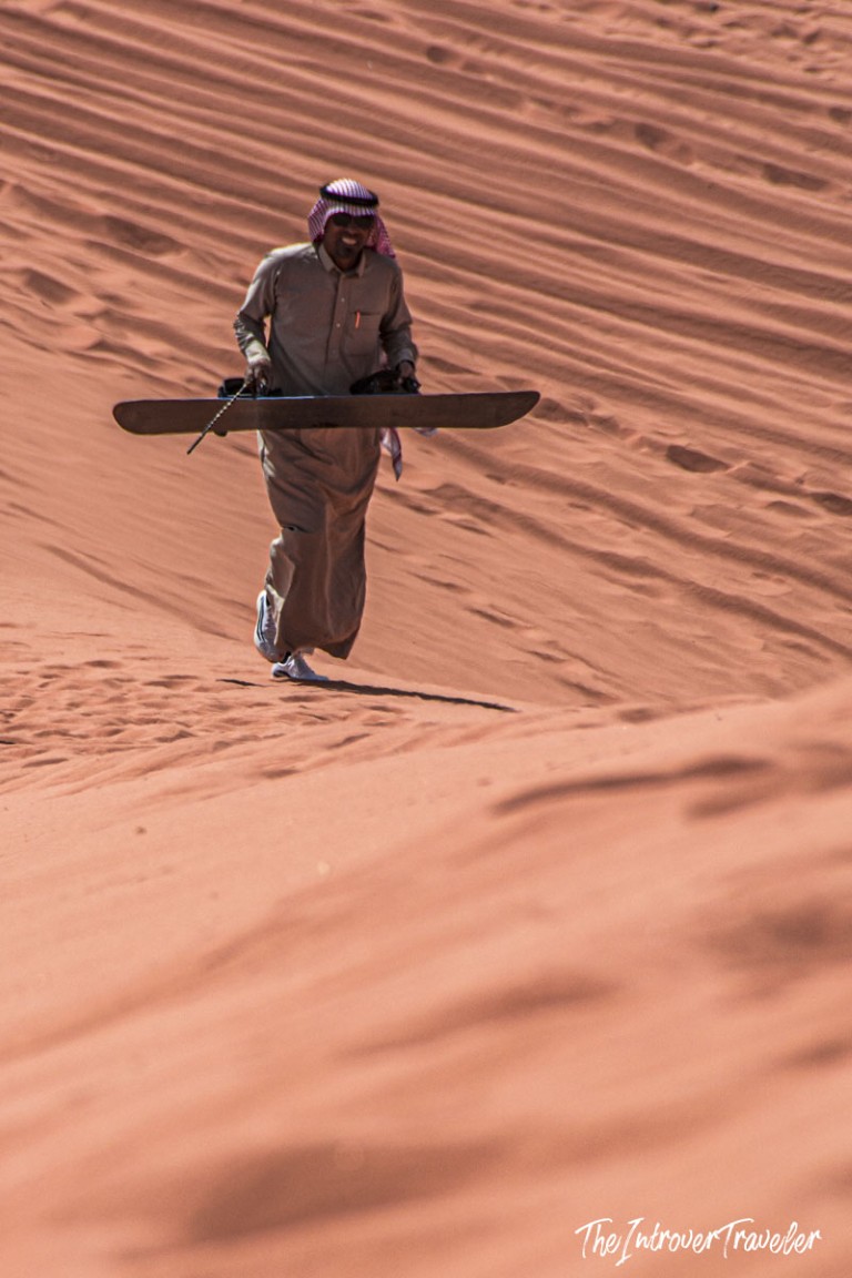 Man in traditional Jordanian dress carrying a sandboard up a red dune in Wadi Rum desert