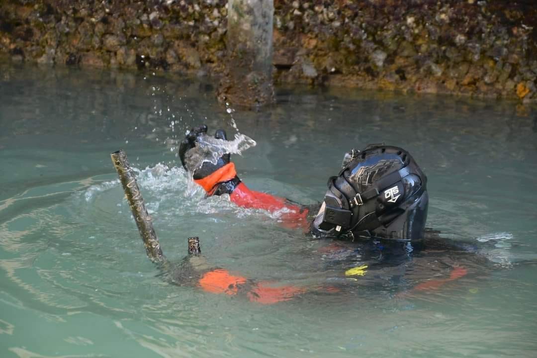 Diver in OTS helmet and orange drysuit working on a submerged wooden gondola mooring pole in a Venice canal