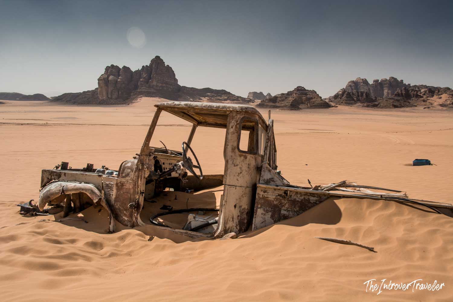 Rusted vehicle wreck half-buried in Wadi Rum sand, with iconic sandstone rock formations behind