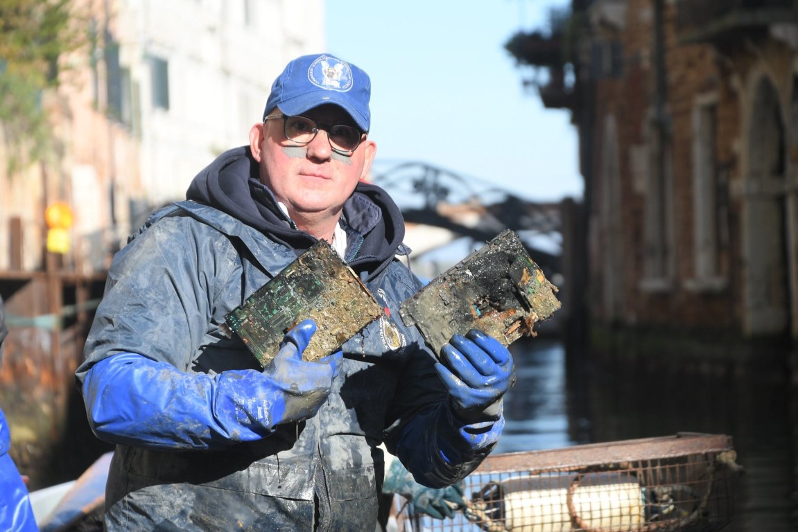 Worker holding mud-covered circuit boards retrieved from a Venice canal during a cleanup dive