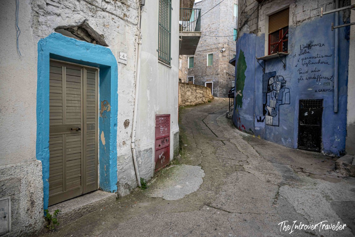 Narrow Orgosolo alley with a Gramsci quote mural on a blue wall beside a turquoise-framed door