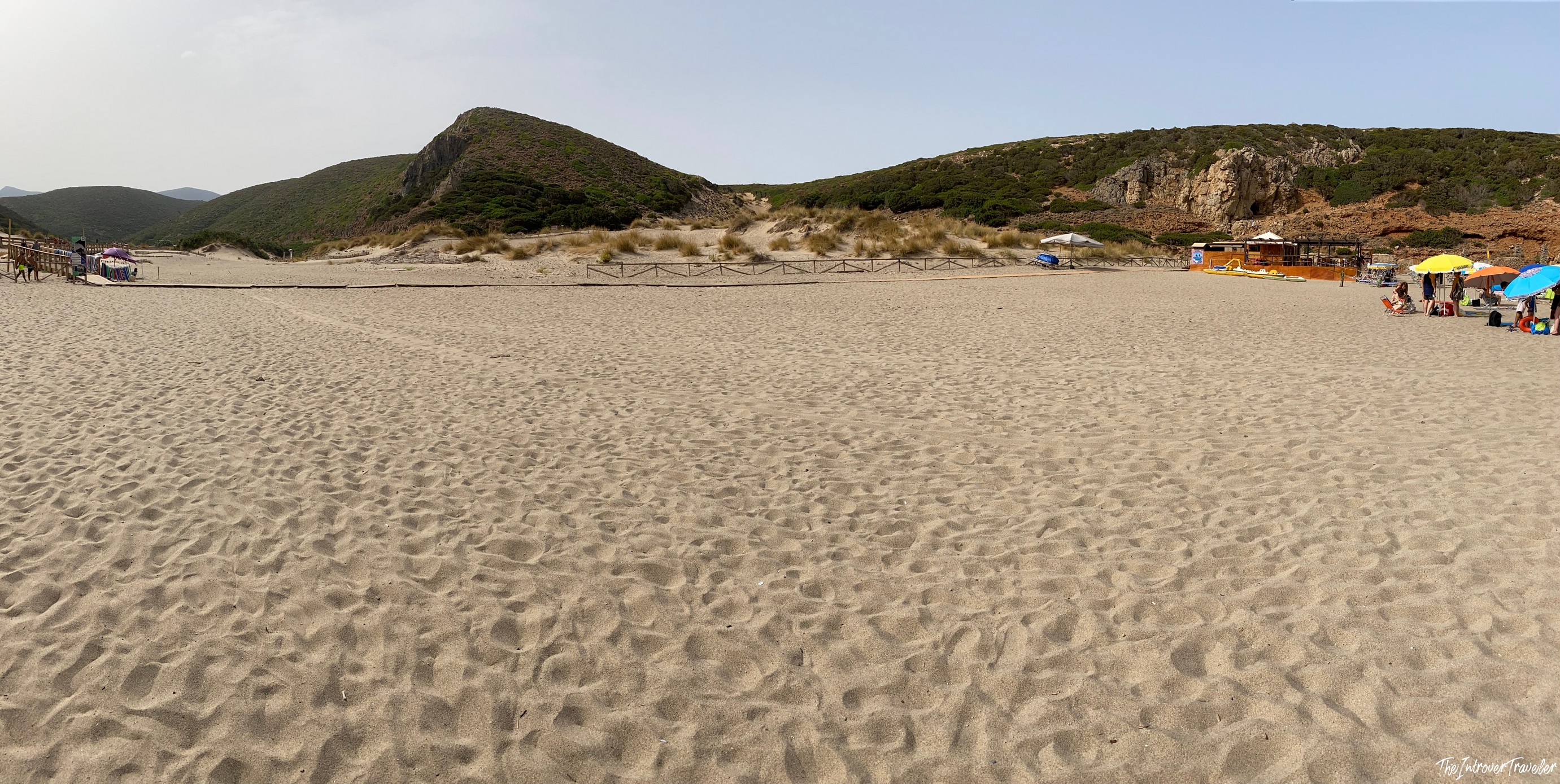 Wide sandy beach in south Sardinia, with grass-covered dunes, rocky hills, and colorful beach umbrellas