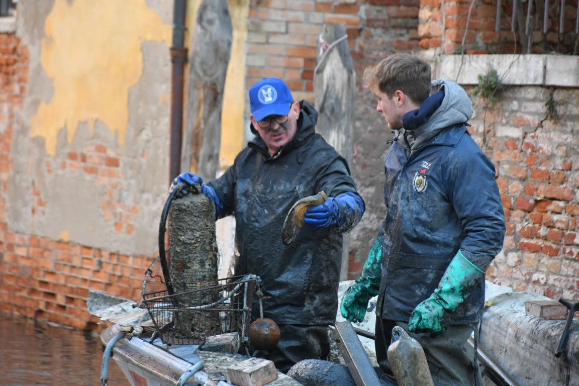 Two workers in muddy wetsuits retrieving debris from a Venice canal on a maintenance boat
