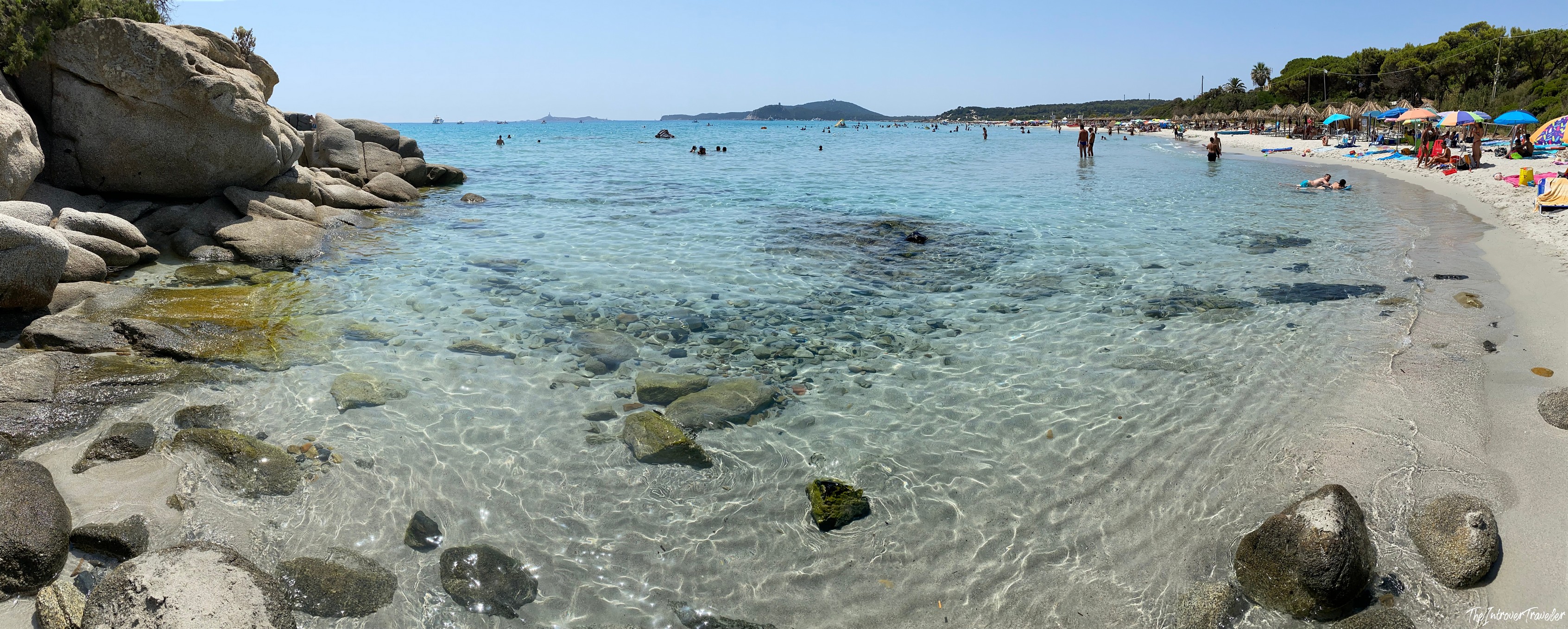 Crystal-clear shallow waters and granite boulders at a south Sardinia beach, with sunbathers and thatched umbrellas