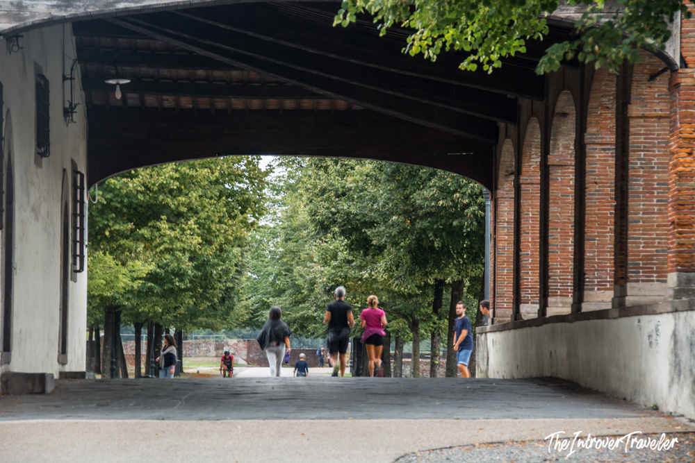 Visitors walking and jogging through a covered gateway in Lucca, Tuscany, along the tree-lined city walls path