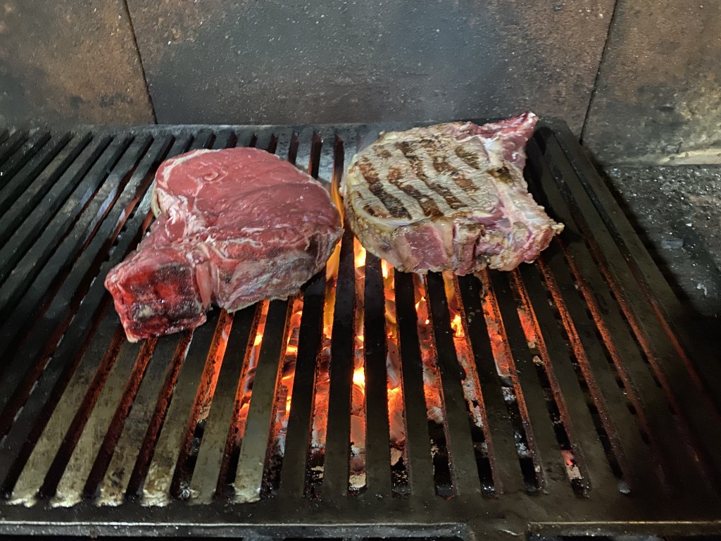 Two thick bistecca steaks grilling over open flames at Taverna del Vecchio Borgo, Massa Marittima