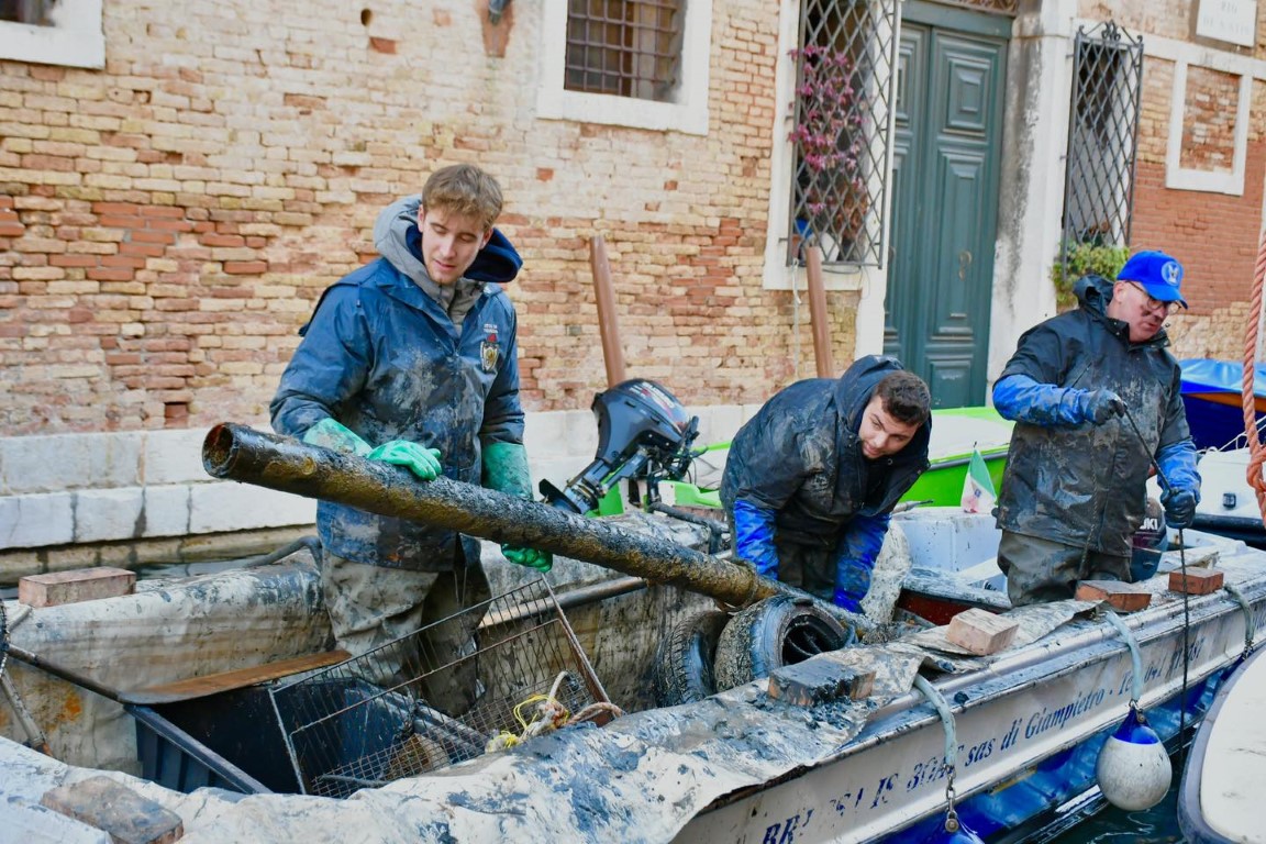 Workers dredging mud and debris from a Venice canal, hauling salvaged pipes onto a work boat