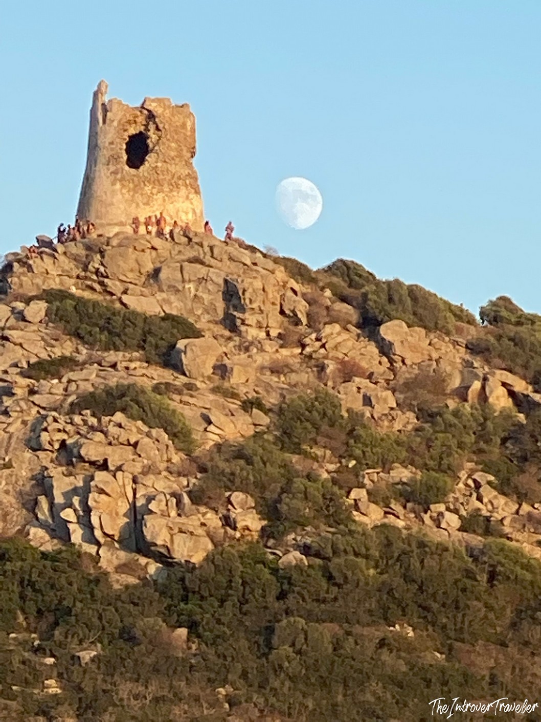 Ancient Sardinian watchtower atop rocky hilltop at golden hour, with full moon rising beside it