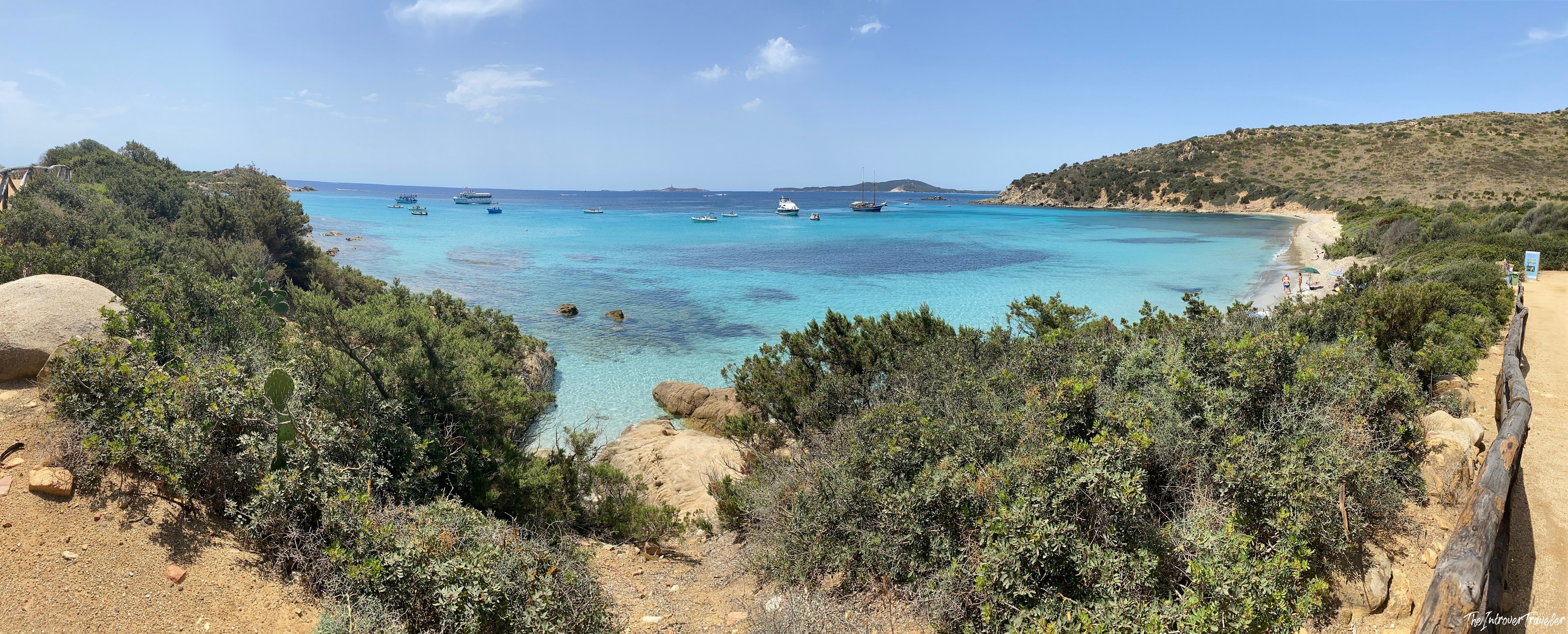 Panoramic view of a turquoise cove in south Sardinia, with anchored boats, scrubland, and a white sand beach