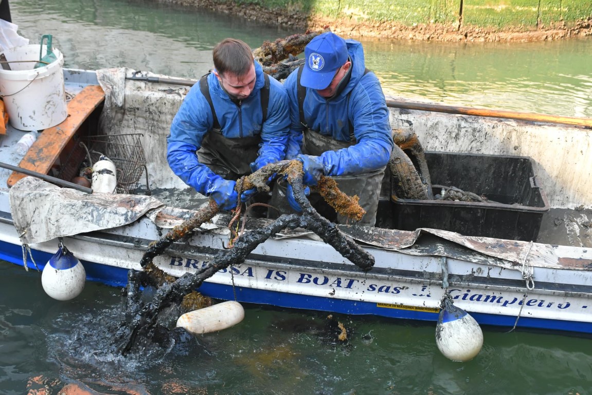 Two Venice canal divers hauling a mud-encrusted metal object from a working boat on a Venetian waterway