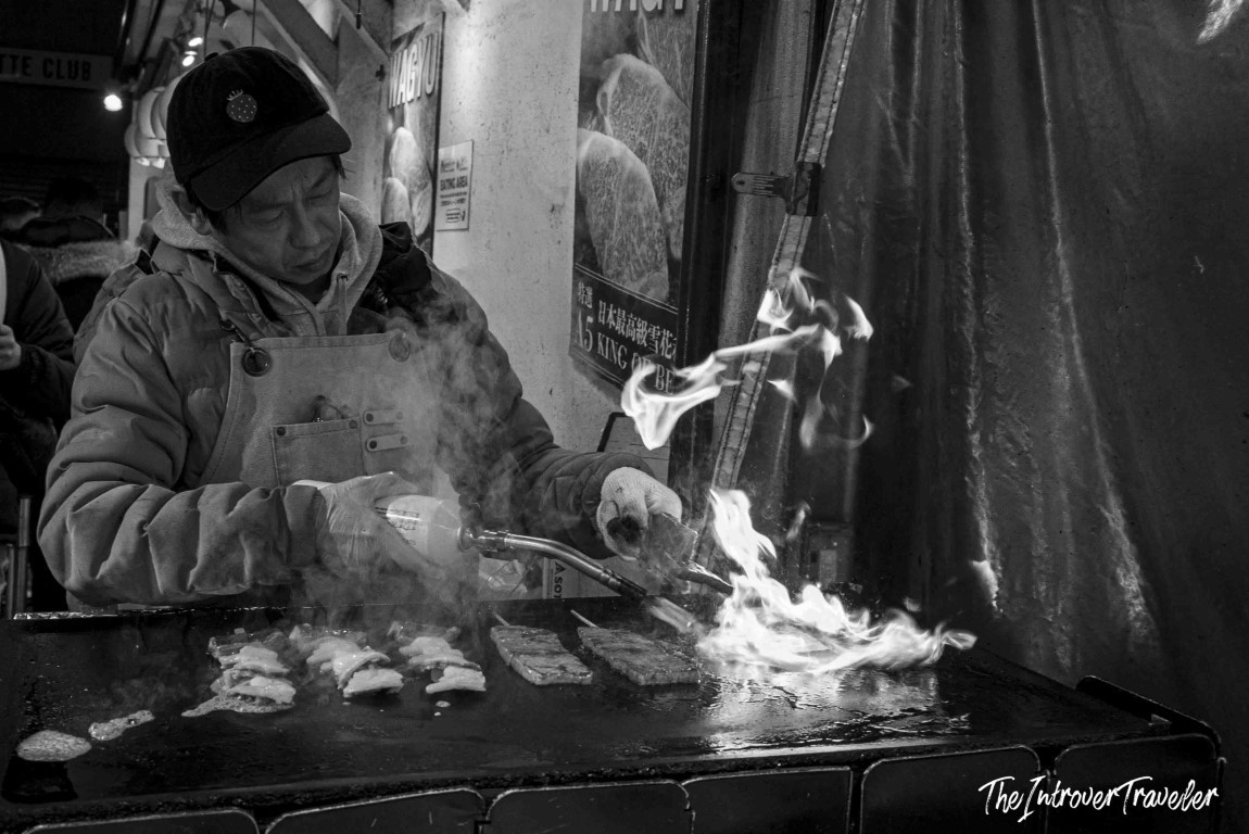 Vendor flame-searing wagyu beef skewers at a bustling Japanese fish market street stall
