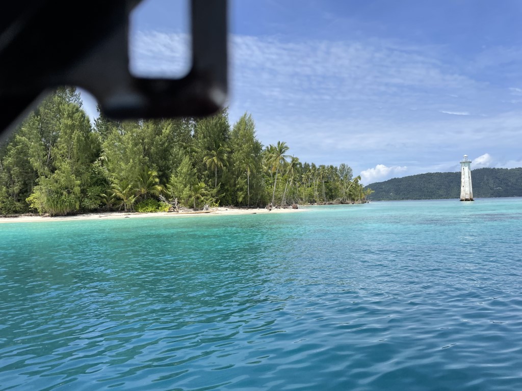 Boat view of a tropical Raja Ampat island, turquoise waters, palm trees, and a small lighthouse