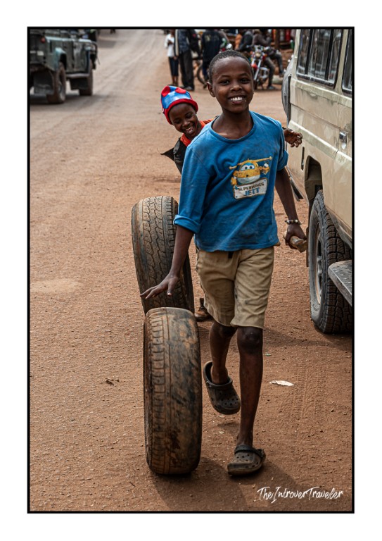 Smiling Tanzanian boy rolling a tire along a dusty village road, capturing everyday local life