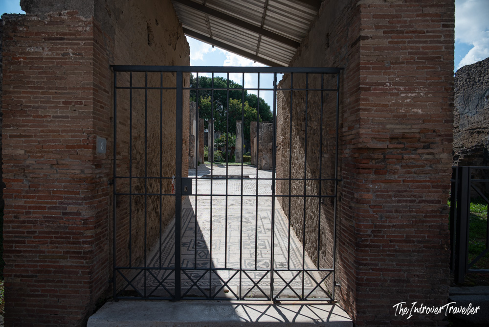 Iron gate at a Pompeii ruins entrance, revealing ancient Roman mosaic floors beyond the brick doorway