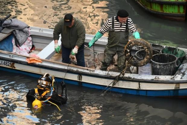 Venice canal diver surfacing beside a small fishing boat, handing up salvaged debris to two workers