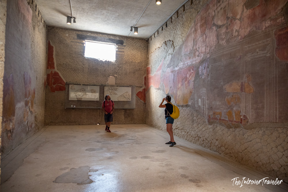 Two visitors photographing faded Roman frescoes inside a preserved room at Herculaneum excavations
