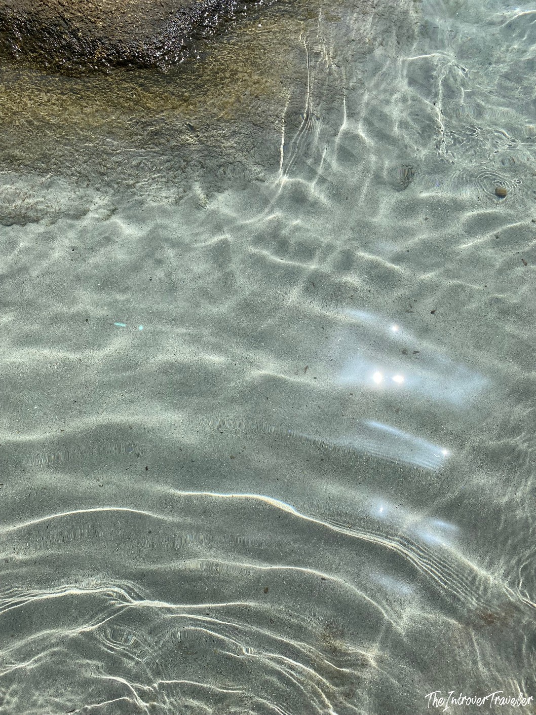 Crystal-clear shallow water over rippled sandy seafloor at a south Sardinia beach, with sunlight glinting off the surface