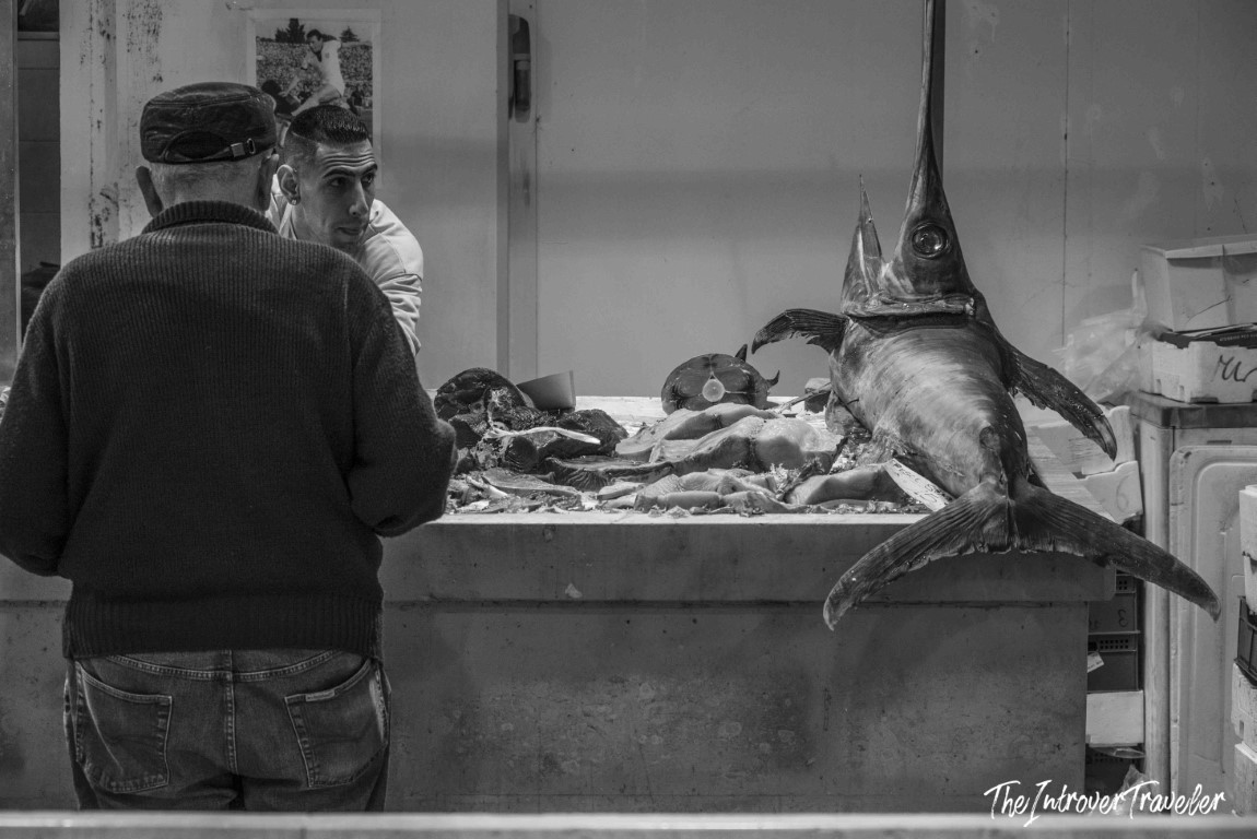 Whole swordfish displayed on ice at a coastal fish market stall, vendor serving a customer